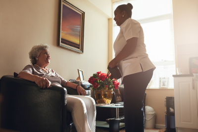 senior woman sitting on a chair at home with female caregiver standing by